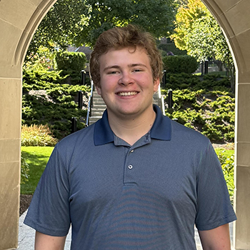 A young person wearing a blue polo shirt stands smiling under a stone archway with greenery and stairs in the background.