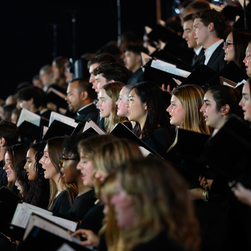 A large choir of men and women sings together while holding sheet music, dressed in black attire, focused and illuminated on stage.