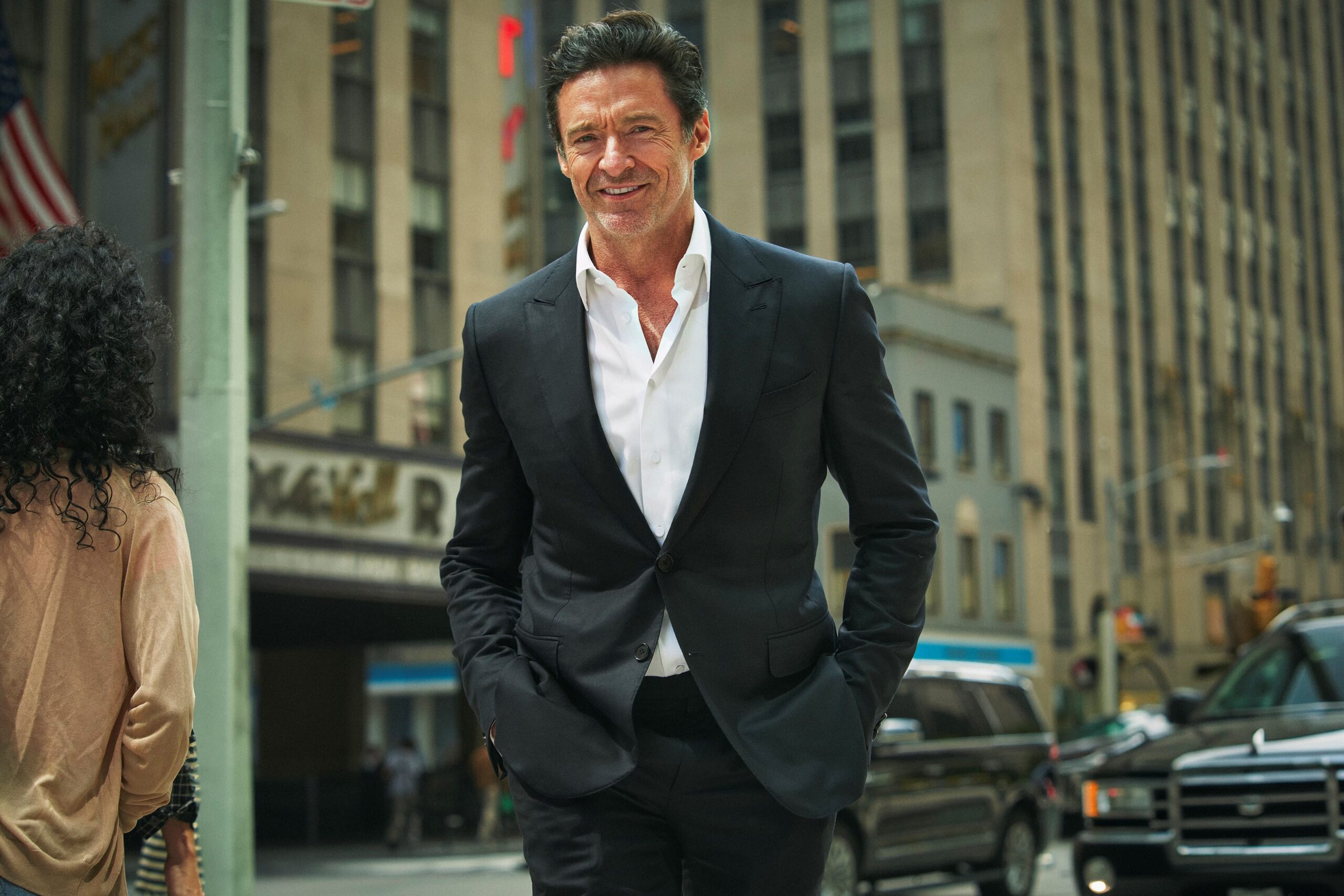 A man in a black suit and white shirt smiles while walking on a city street, with tall buildings, cars, and another person visible in the background.